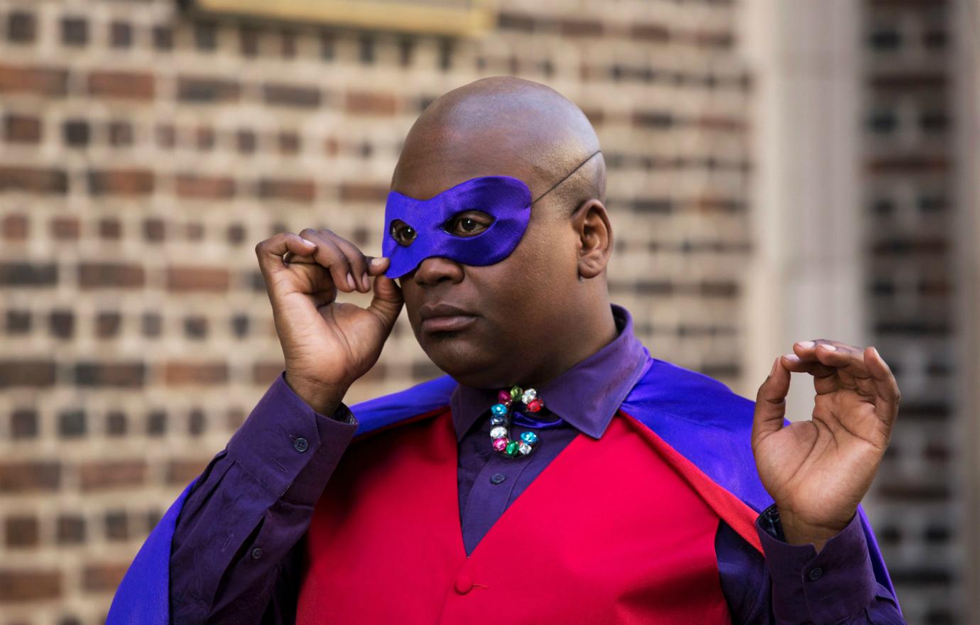 Tituss Burgess as Titus Andromedon wears a red jacket, purple cape, and purple eye covering.