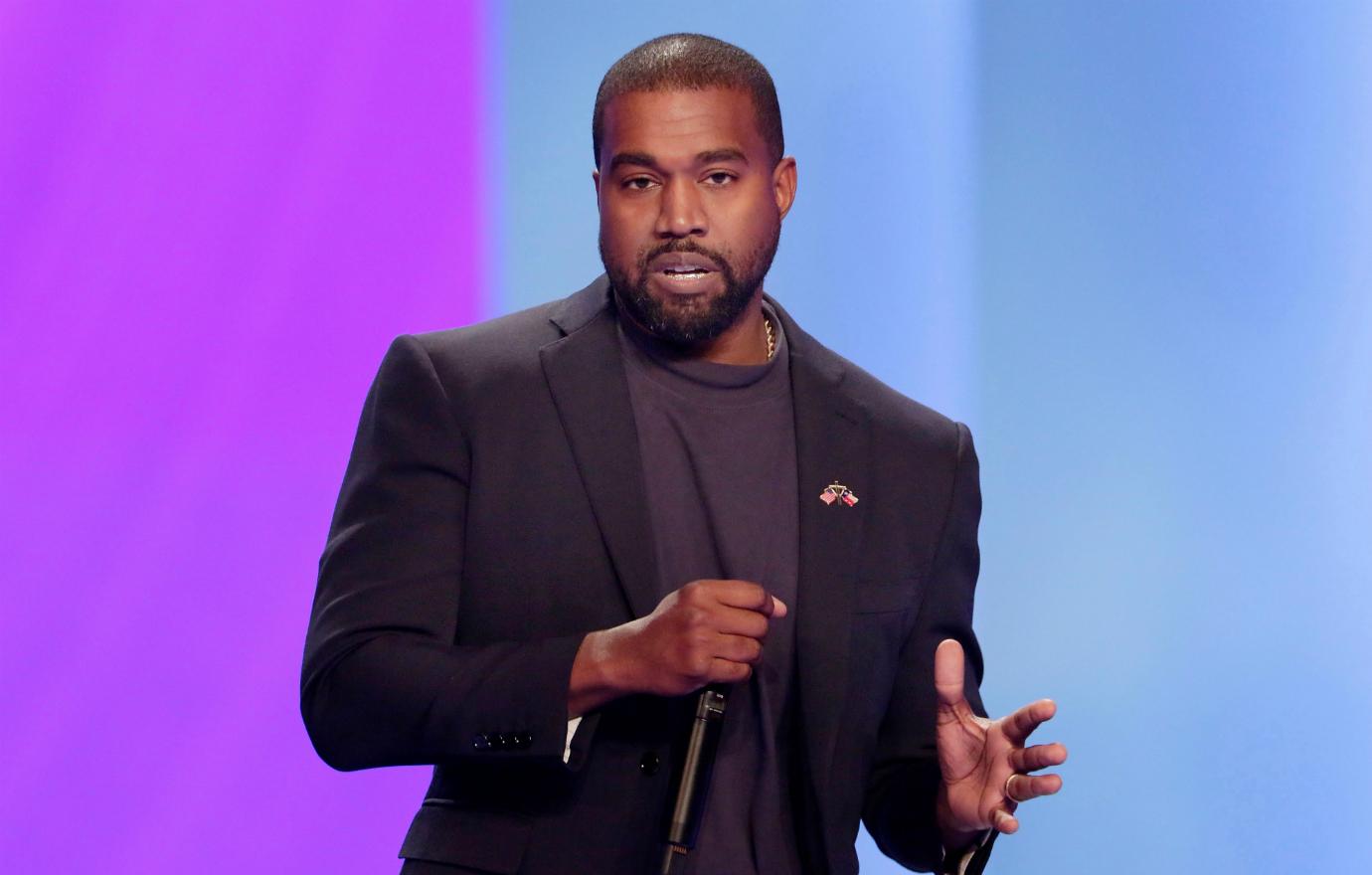 Kanye West, on stage and holding a mic, wears a dark jacket over a dark top.