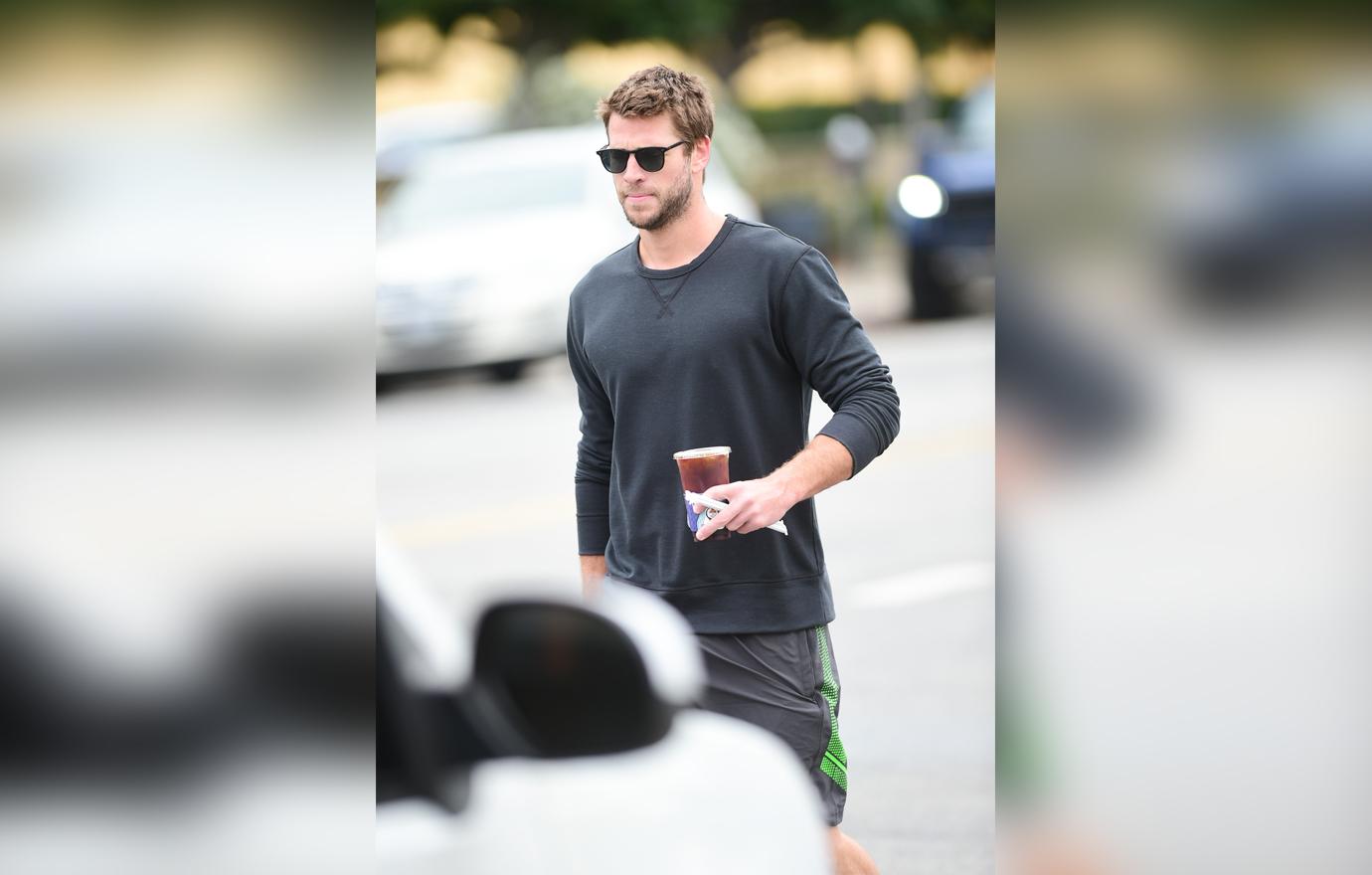 Liam Hemsworth Walking TO His Car Holding An Iced Coffee Looking Serious Wearing Black Sunglasses Shorts and a Long Sleeve Tee