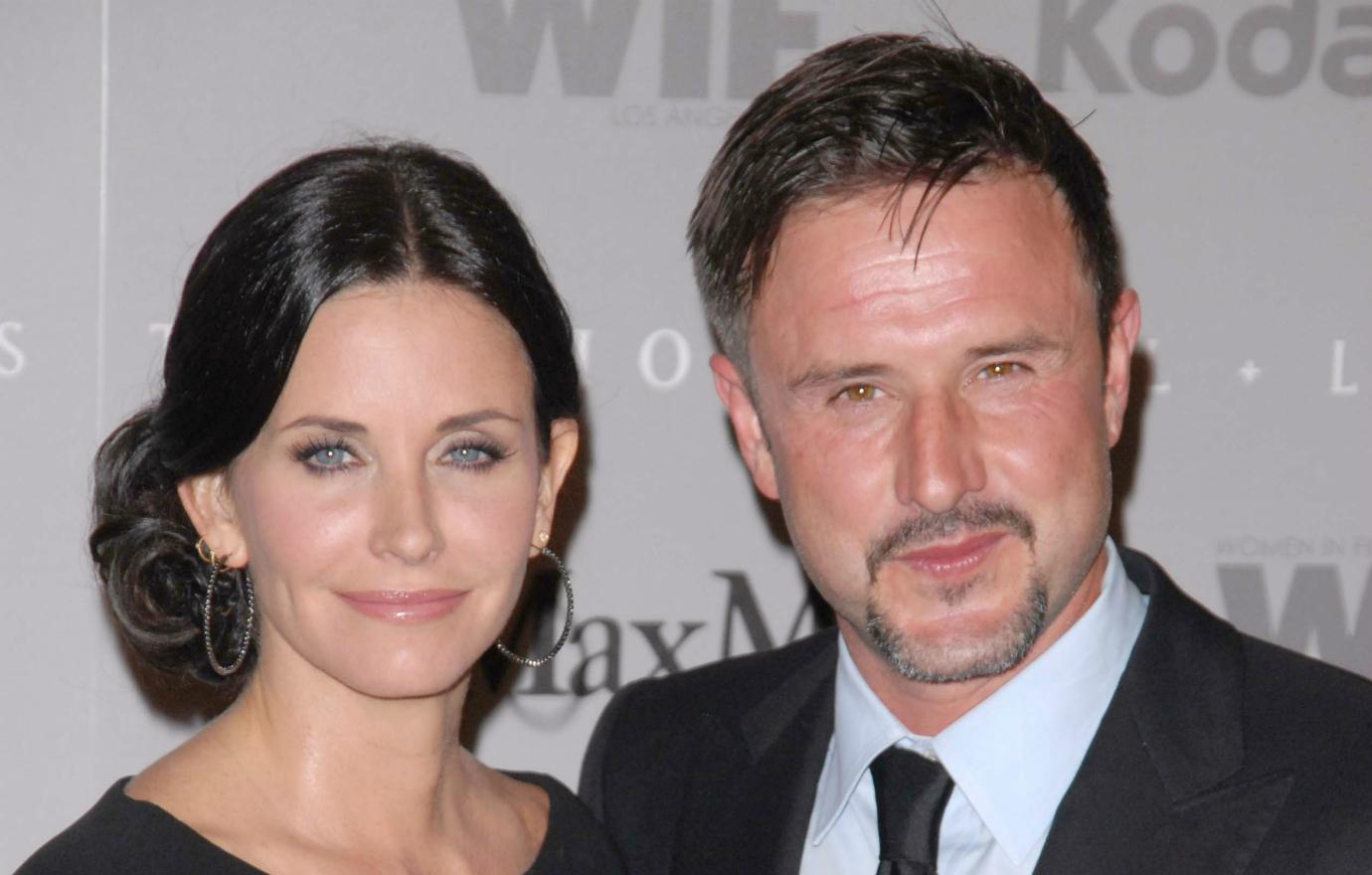 Courtney Cox, in a black dress, stands next to David Arquette who wears a dark jacket, black tie, and blue shirt.