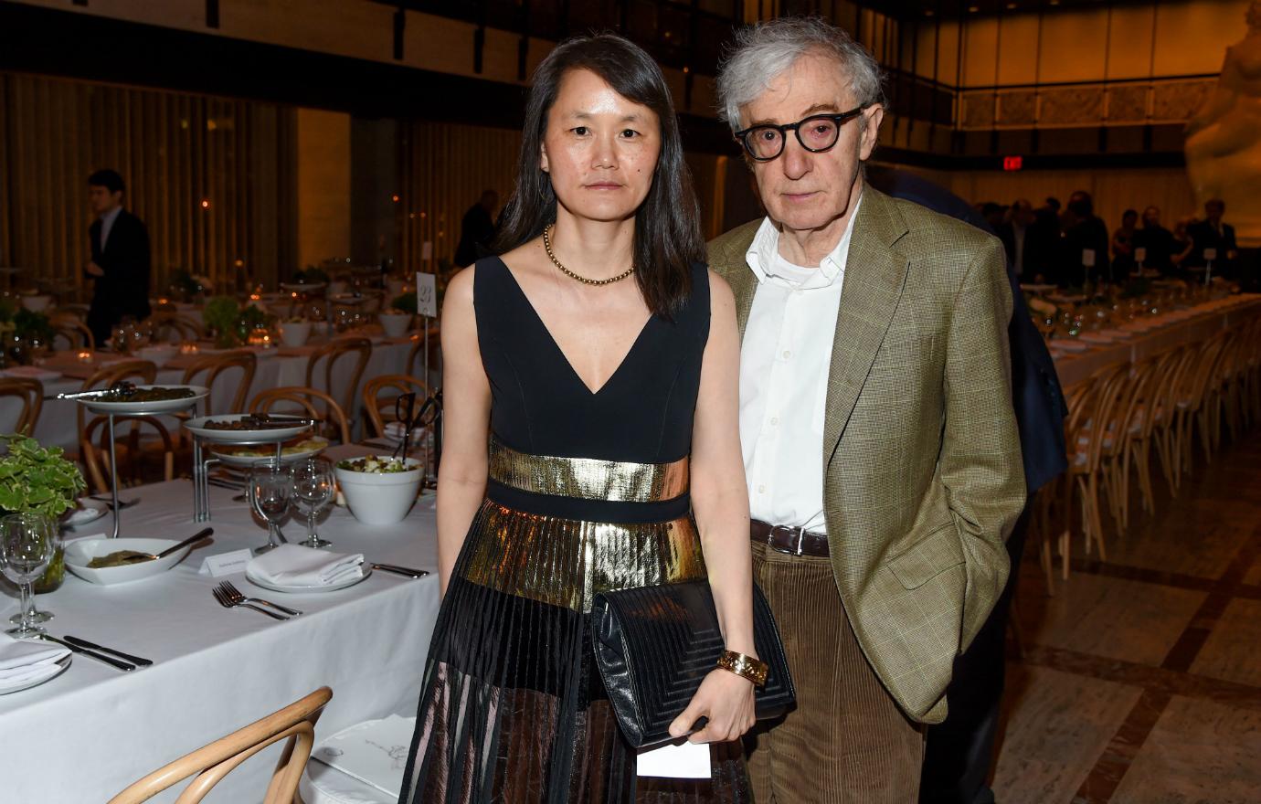 Soon-Yi Previn wears a black dress with gold accents and stands next to Woody Allen who wears beige pants, a white shirt, and olve jacket.