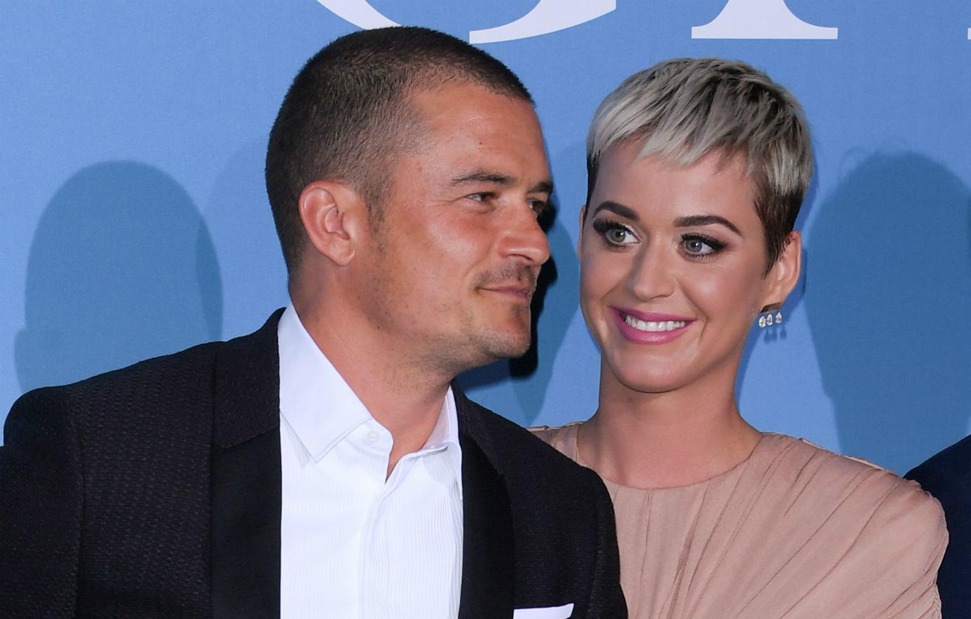 Orlando Bloom, in a black suit and white shirt, stands next to Katy Perry, who wears a pink dress.