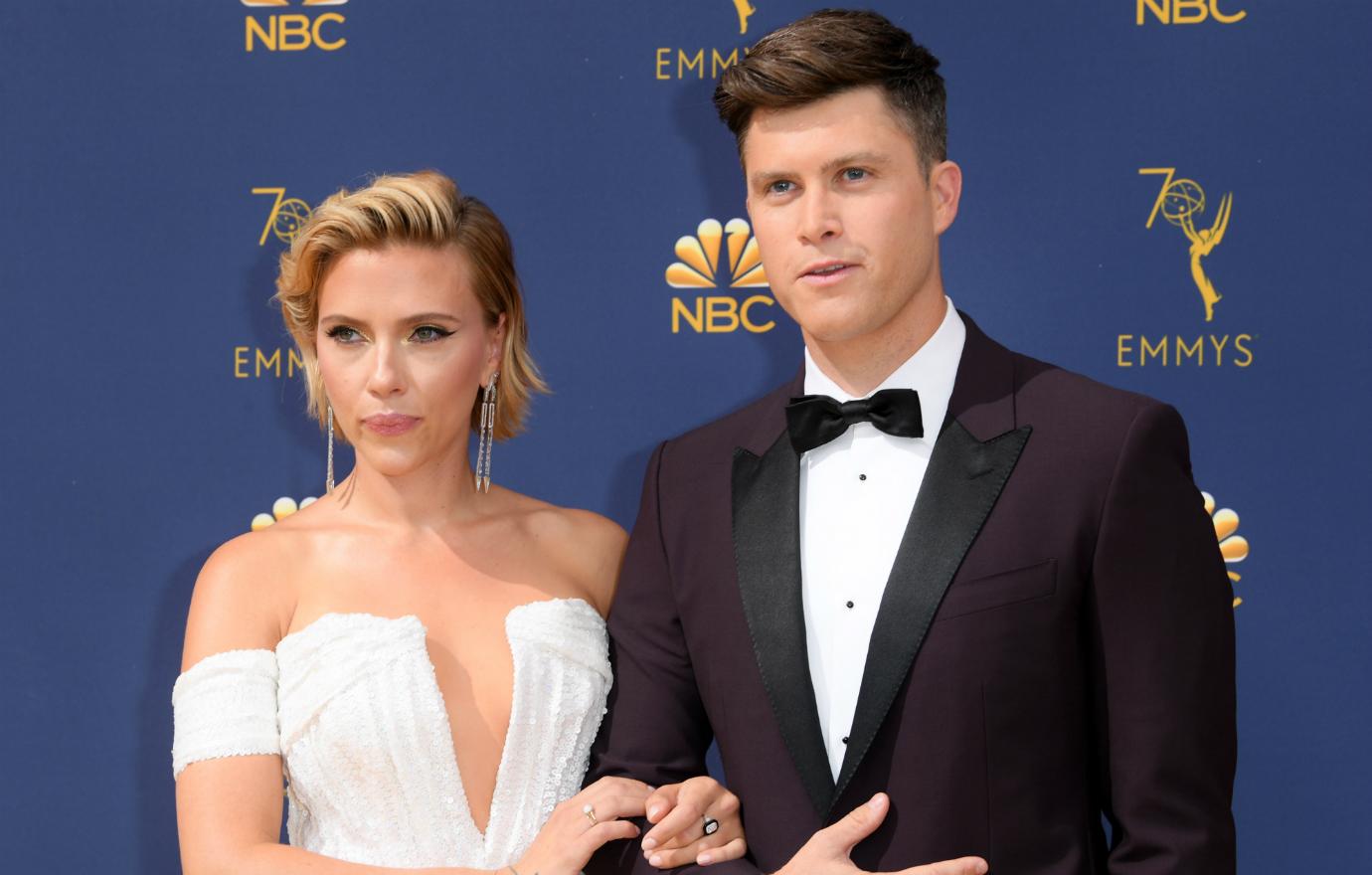 ScarJo, in a white dress, stands on the red carpet with Colin Jost who wears a black tux.