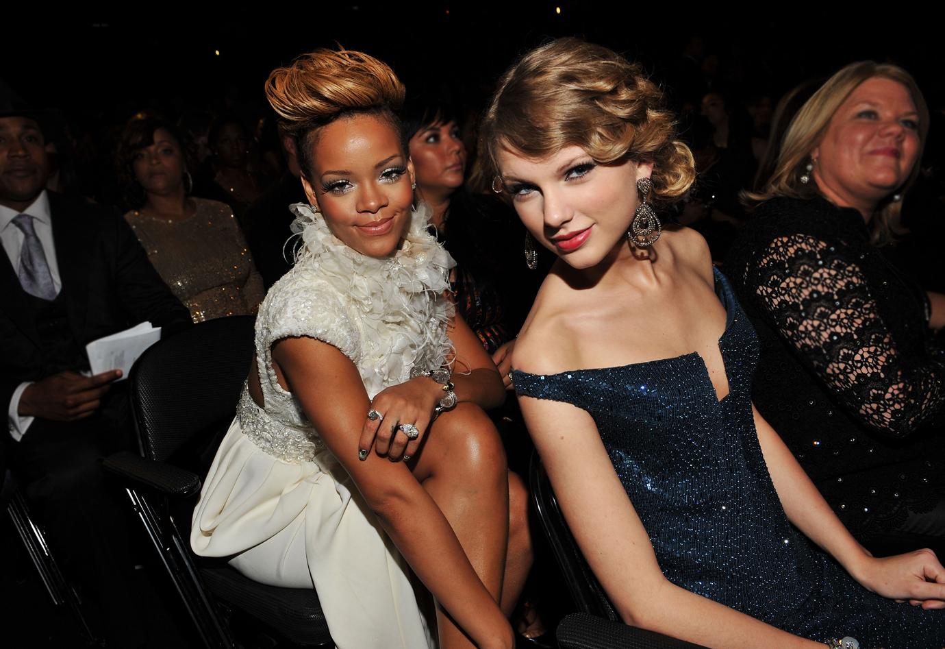 Rihanna Wearing White Gown Sits Behind Taylor Swift Wearing Navy Dress As They Both Smile At The Camera