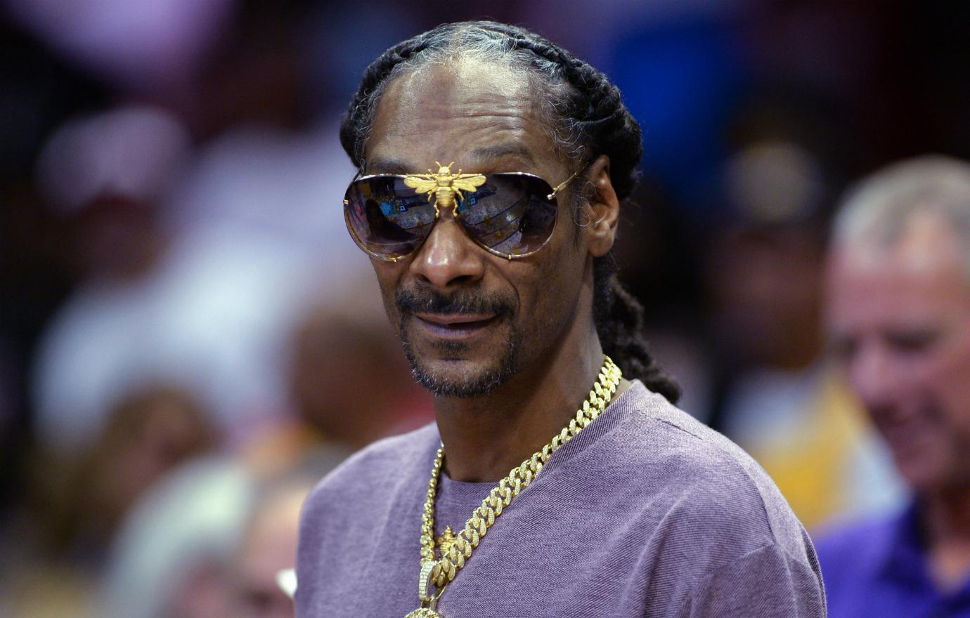 Snoop Dogg wears a purple tshirt and gold chains and sunglasses.