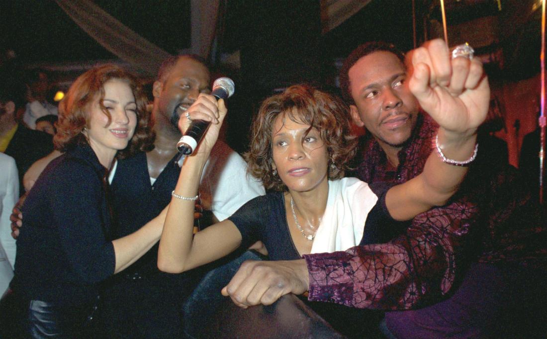 Whitney Houston, in black and clutching a mic, is flanked by Bobby Brown and Gloria Estefan.