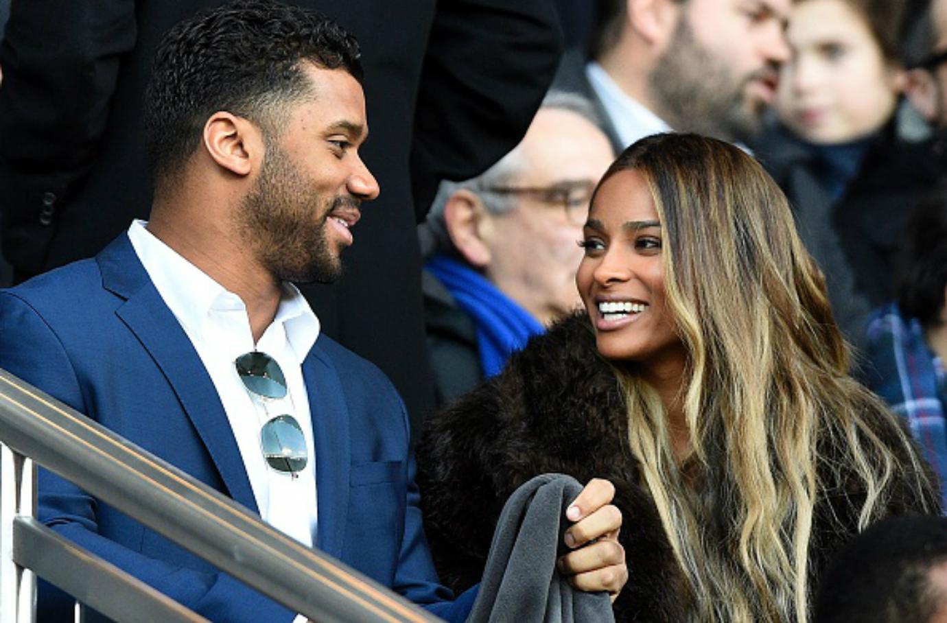 Russel Wilson and Ciara talk while sitting in the stands at a sporting event.