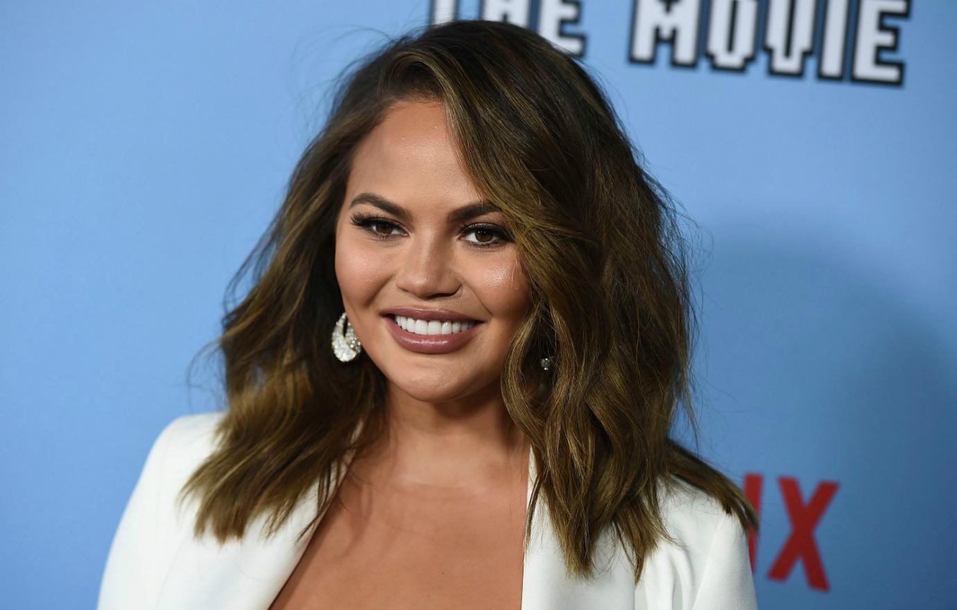 Chrissy Teigan, in a white suit, smiles on the red carpet.