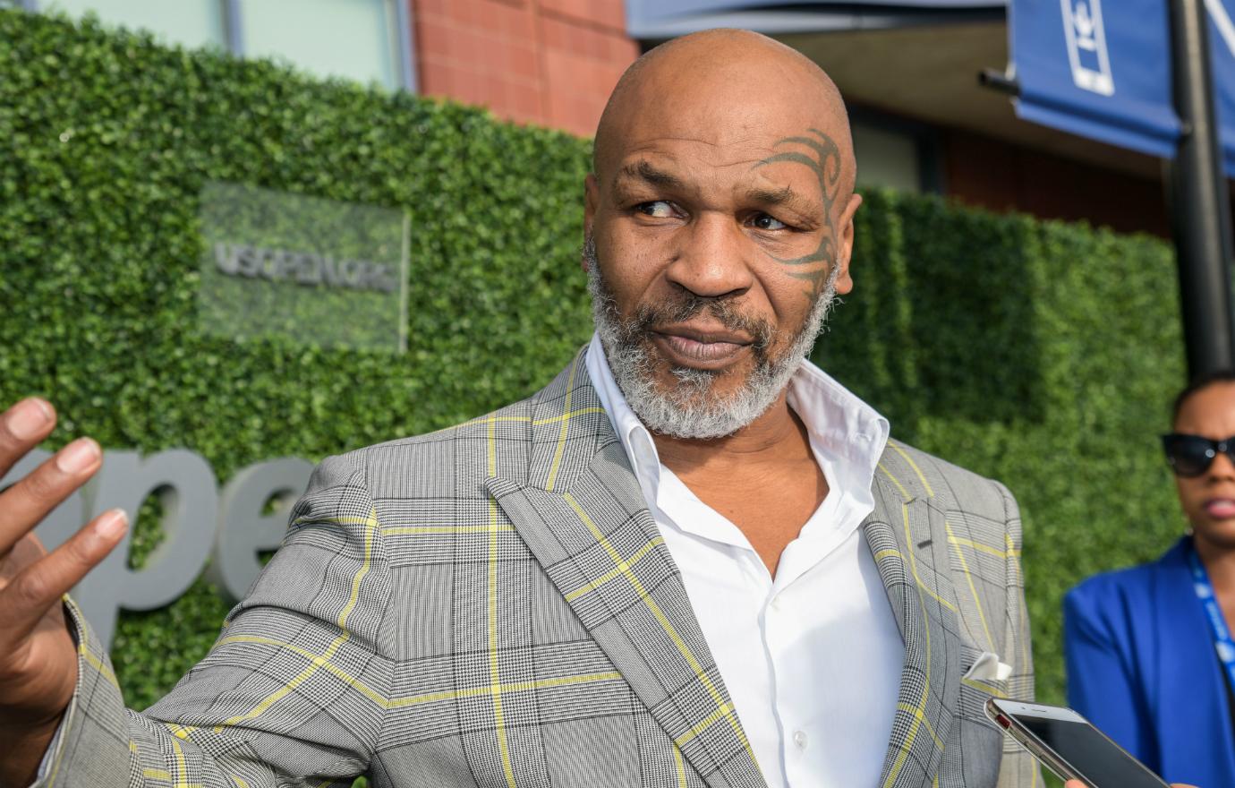 Mike Tyson, wears a checked grey and yellow jacket over a white shirt and stands on the red carpet.