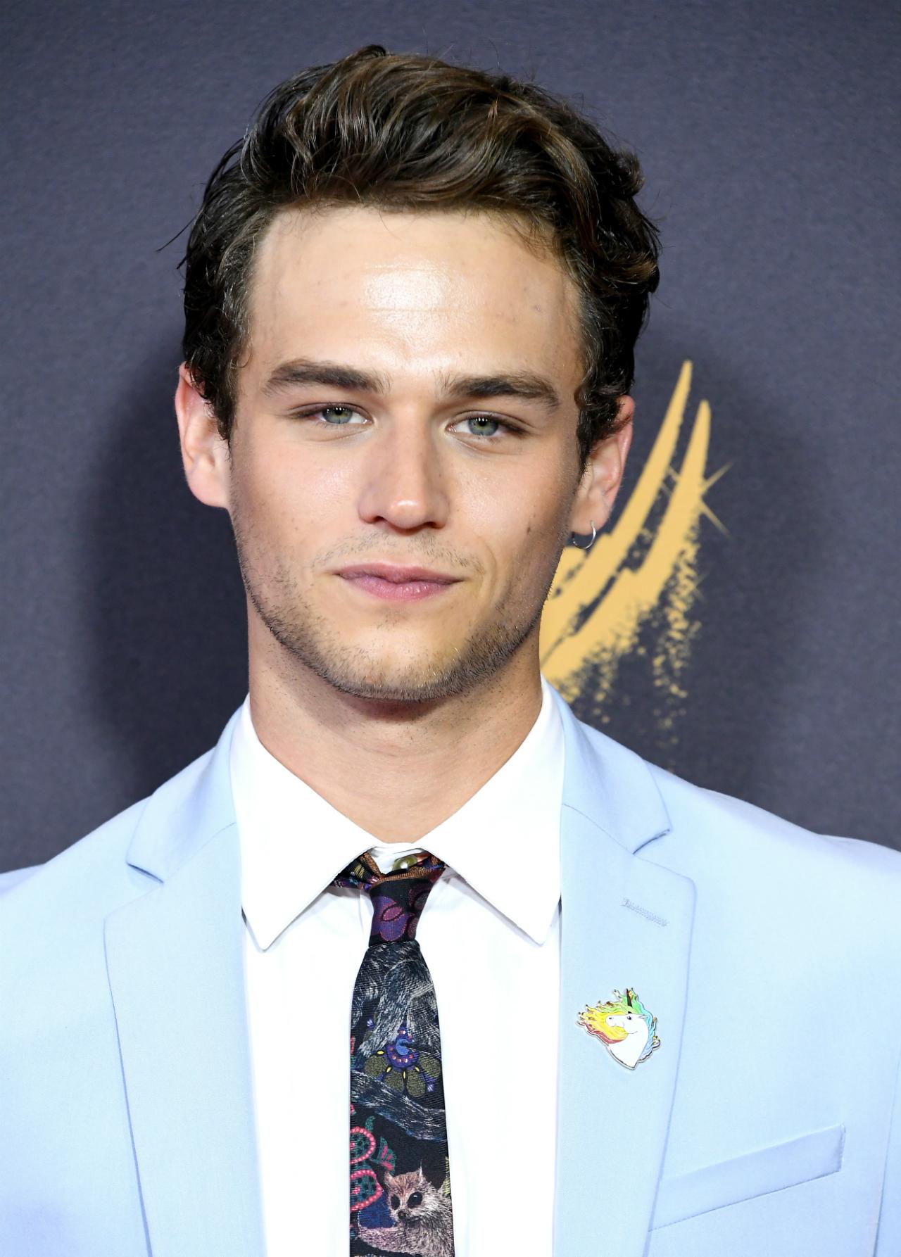 Brandon Flynn wears a light blue suit jacket, white shirt and patterned blue tie, as well as a unicorn rainbow pin on his lapel.