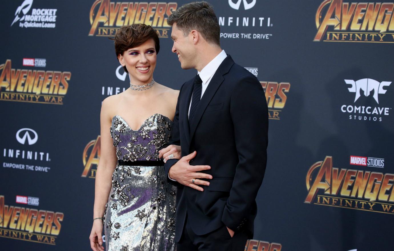 Colin Jost, in a black suit, links arms with Scarlett Johansson who wears a silver dress.