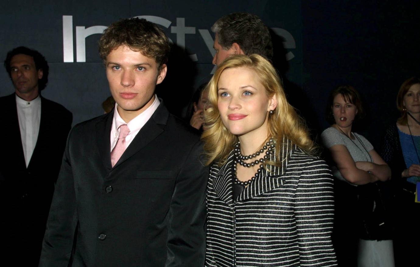 Ryan Phillippe, in a black suit, white shirt, and pink tie, stands on the red carpet next to Reese Witherspoon who wears a black and white striped jacket.