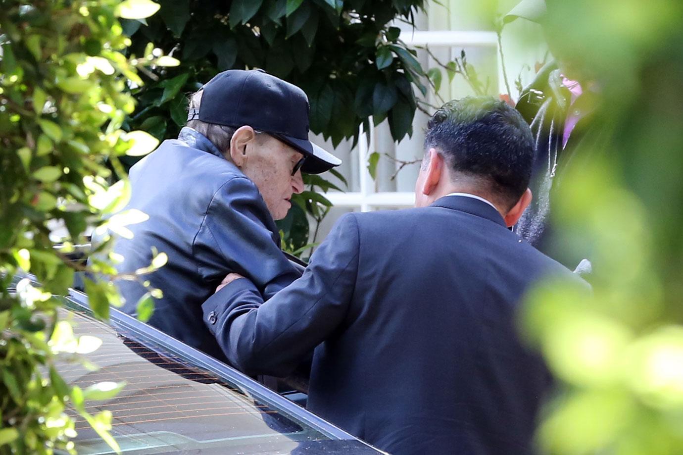 Larry King Wearing Baseball Cap Being Helped Out Of Car