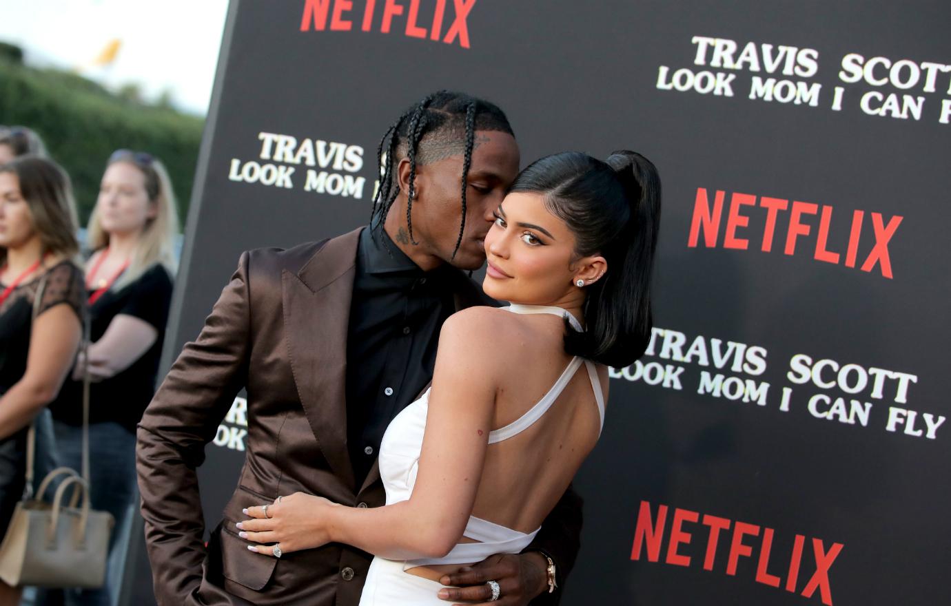 Travis Scott, in a brown jacket and black shirt, kisses Kylie Jenner, who wears a white dress, on the red carpet.