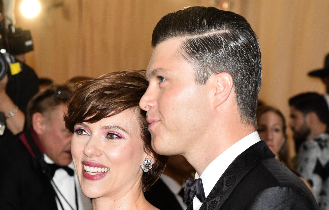 A close up of Colin Jost, in a black suit, standing next to Scarlett Johansson on the red carpet.