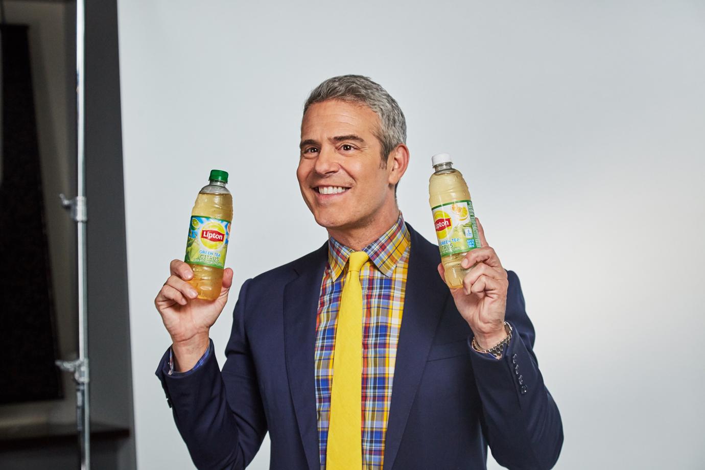 Andy Cohen Wearing Yellow Tie Plaid Shirt and Navy Blazer Smiling In Front of White Backdrop and Posing With a Bottle in Each Hand of Lipton Tea