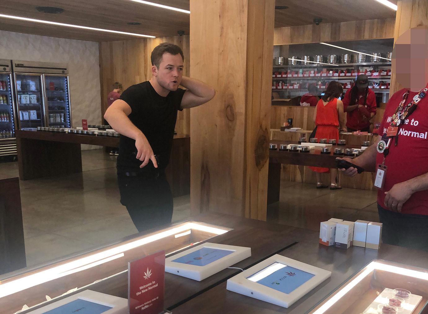 Taron Egerton Wearing Black T-Shirt and Black Jeans Pointing at Display Inside a Medical Marijuana Store