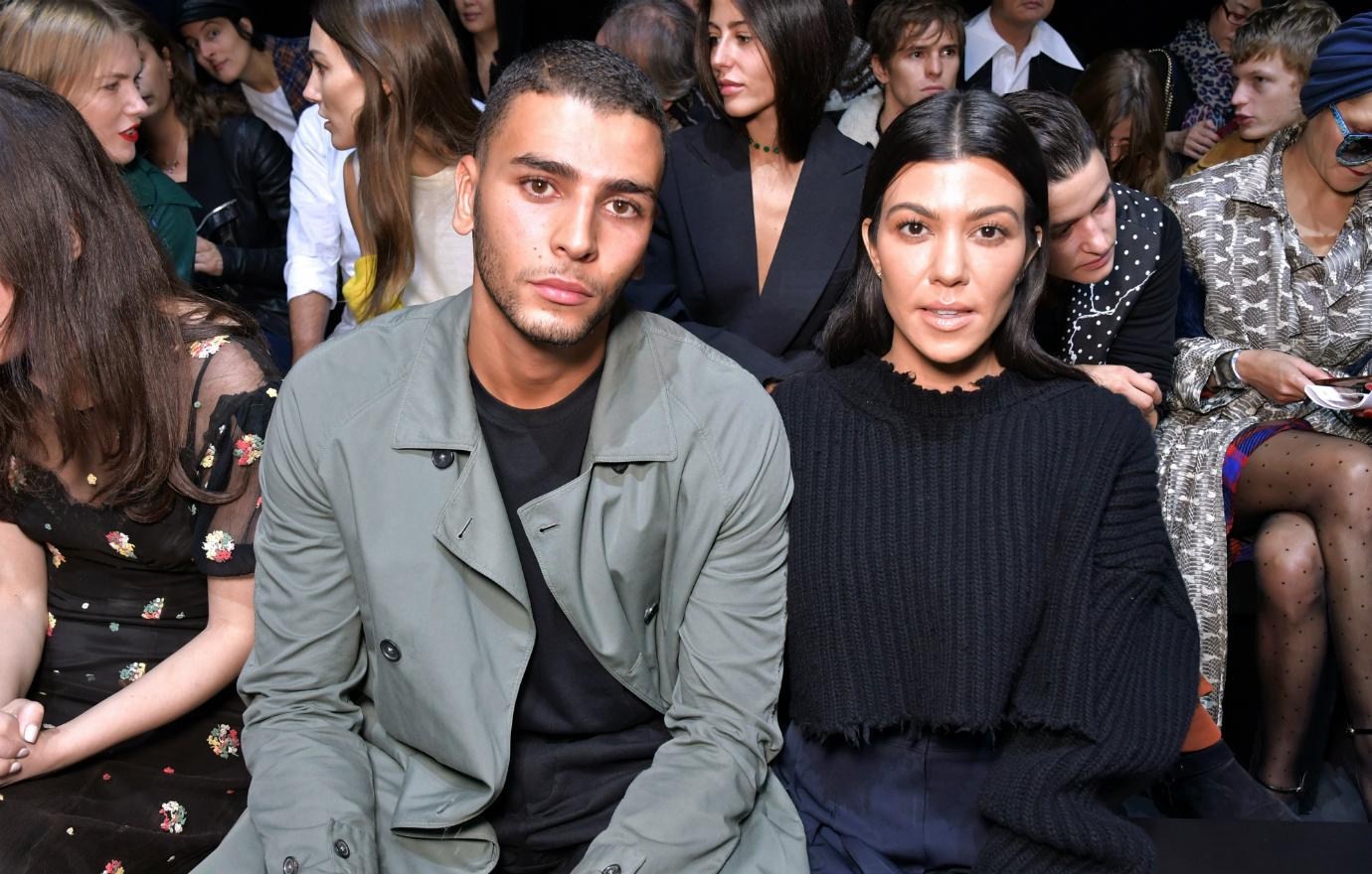 Kourtney Kardashian is all in black and sits next to Younes Bendjima who wears a grey jacket and black top.