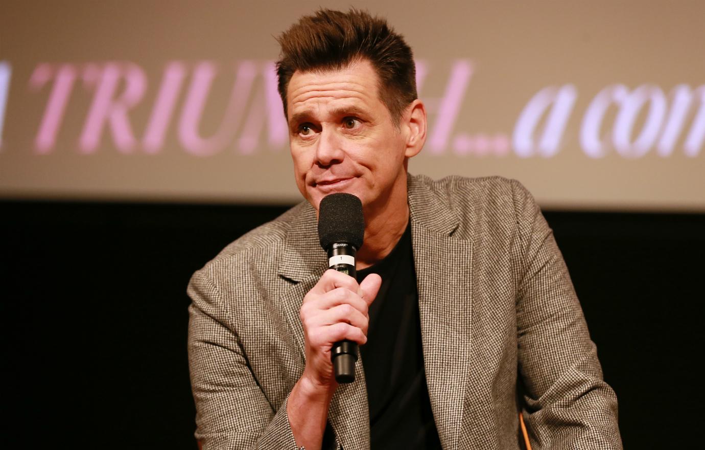 Jim Carrey in a brown jacket and black sweater holds a microphone and talks to an audience at a screening for Showtime’s Kidding.