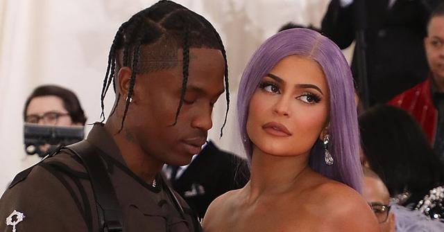 Kylie Jenner wearing a purple dress looking away from Travis scott who wears a faux military harness and brown shirt.