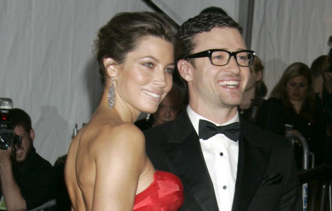 Jessica Biel, in a red gown, stands alongside Justin Timberlake who wears a black tux and white shirt and glasses.