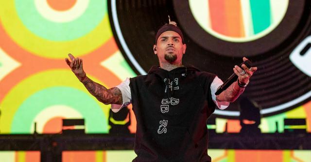 Chris Brown stands on stage, arms outstretched, in a black shirt and blue jeans.