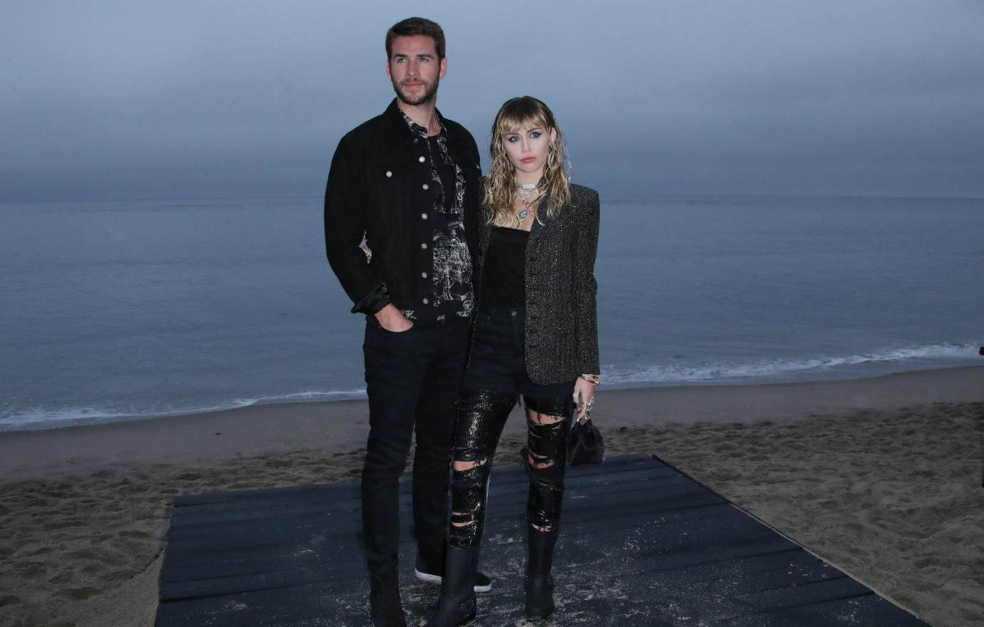 Liam Hemsworth, in a black jacket, black jeans, and black top stands on the beach next to Miley Curys who wears torn black jeans, black top, and a leather jacket.