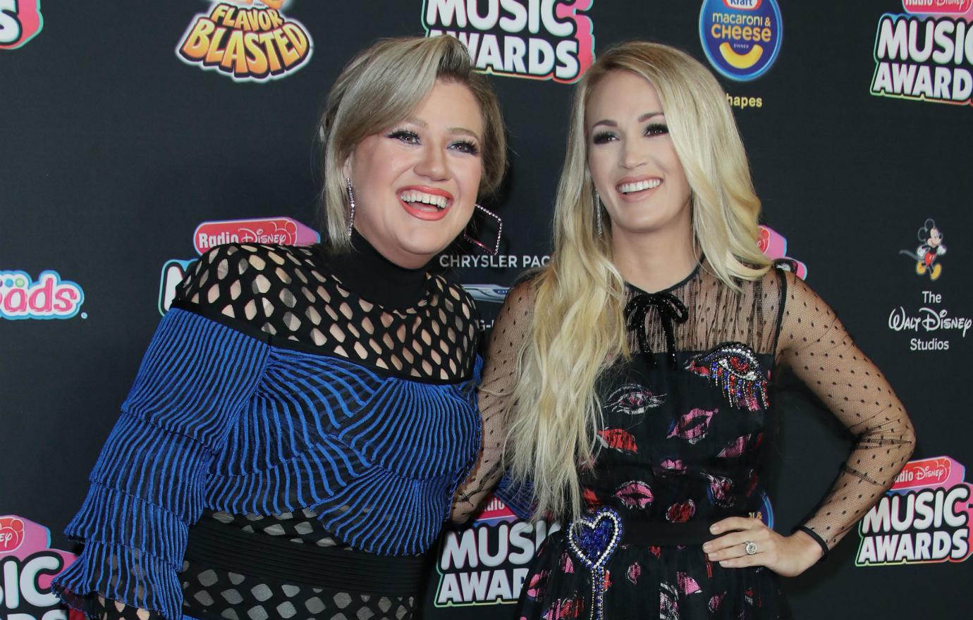 Kelly Clarkson, in a blue/black dress, stands next to Carrie Underwood, in a flowered black dress, on the red carpet.