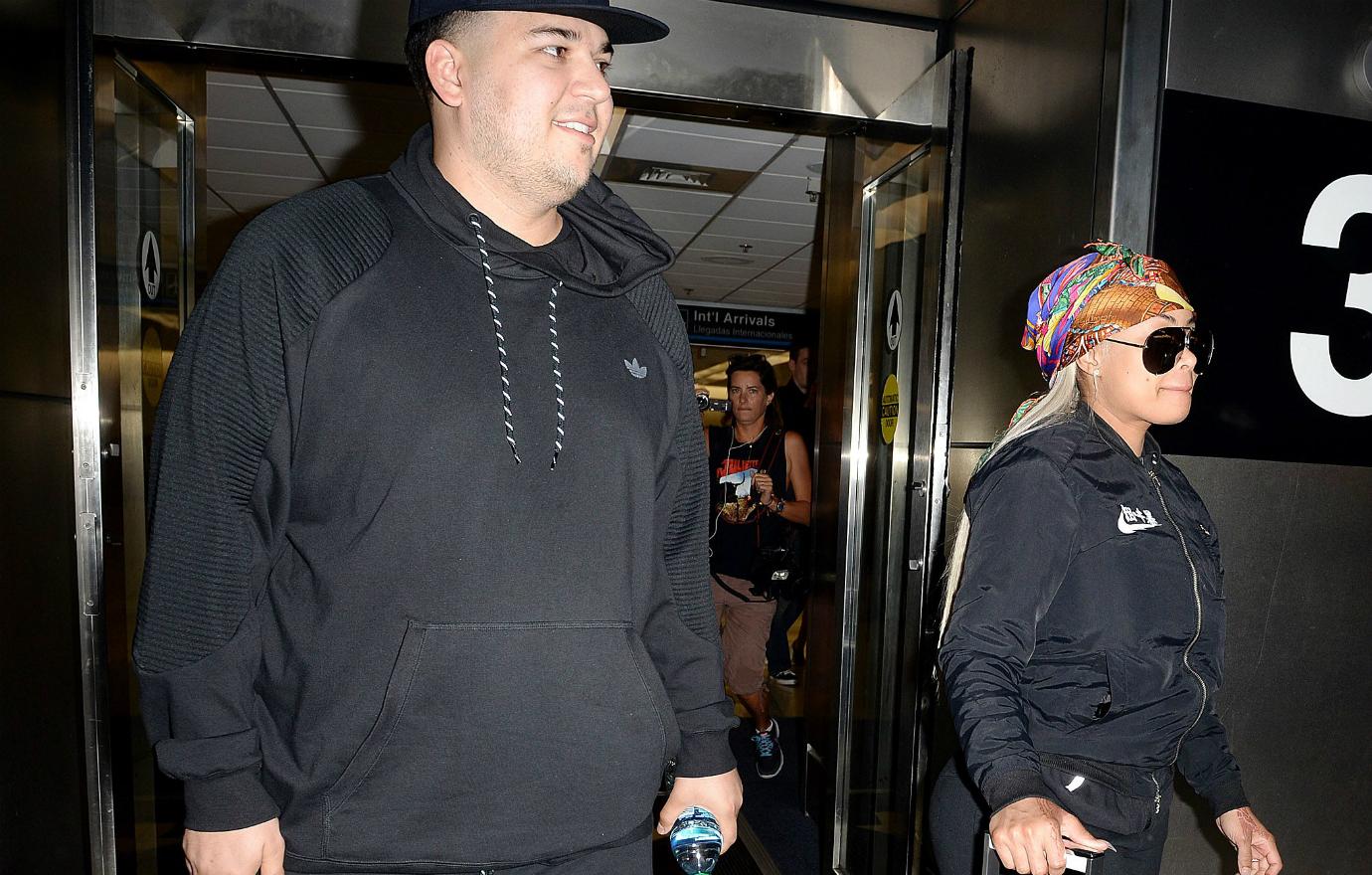 Rob Kardashian, in a black track suit, walks through an airport with Blac Chyna who wears a black track suit, sunglasses, and a bandanna.