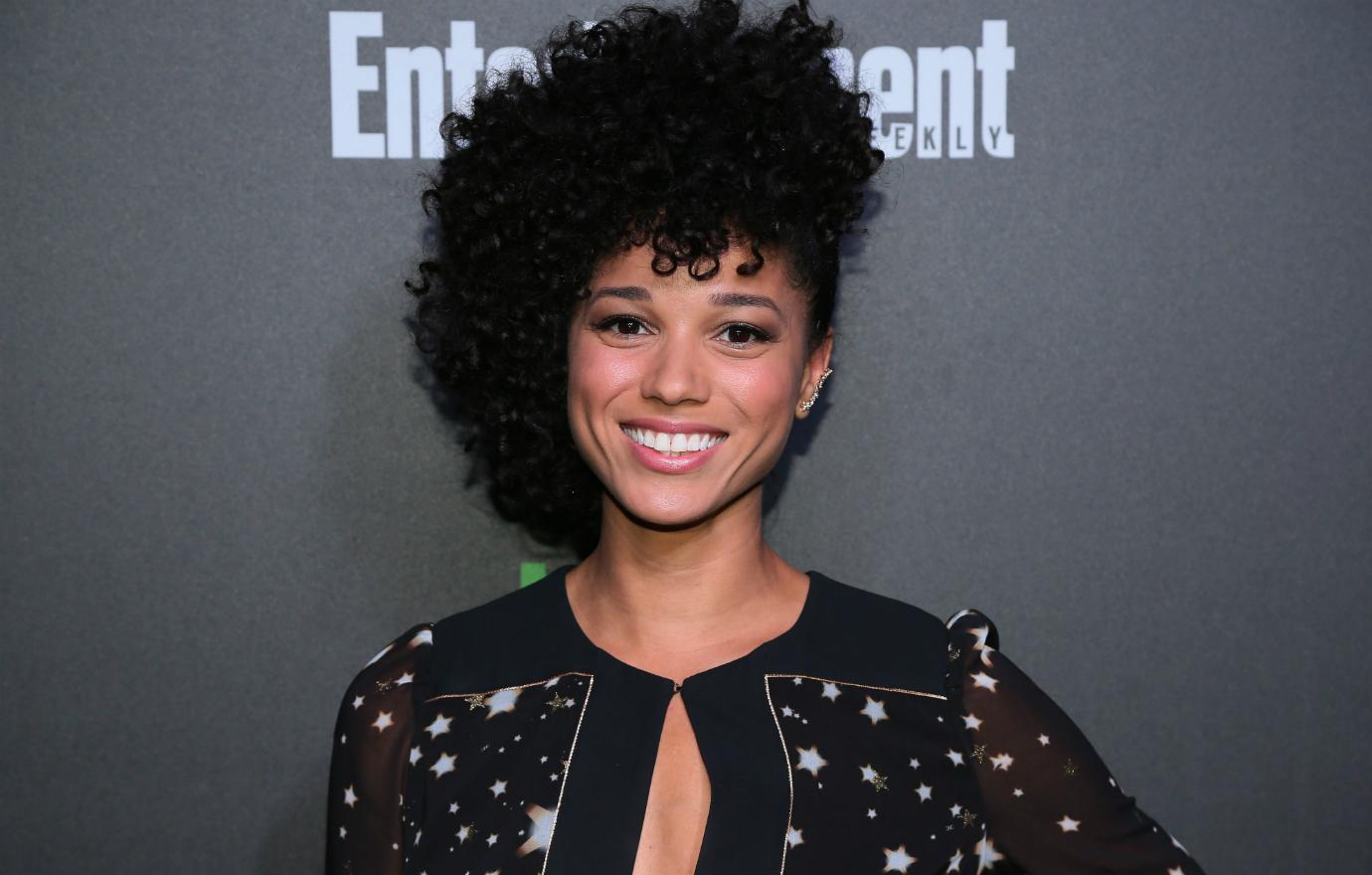 Alisha Wainwright smiles on the red carpet while wearing a black dress with white stars on it.