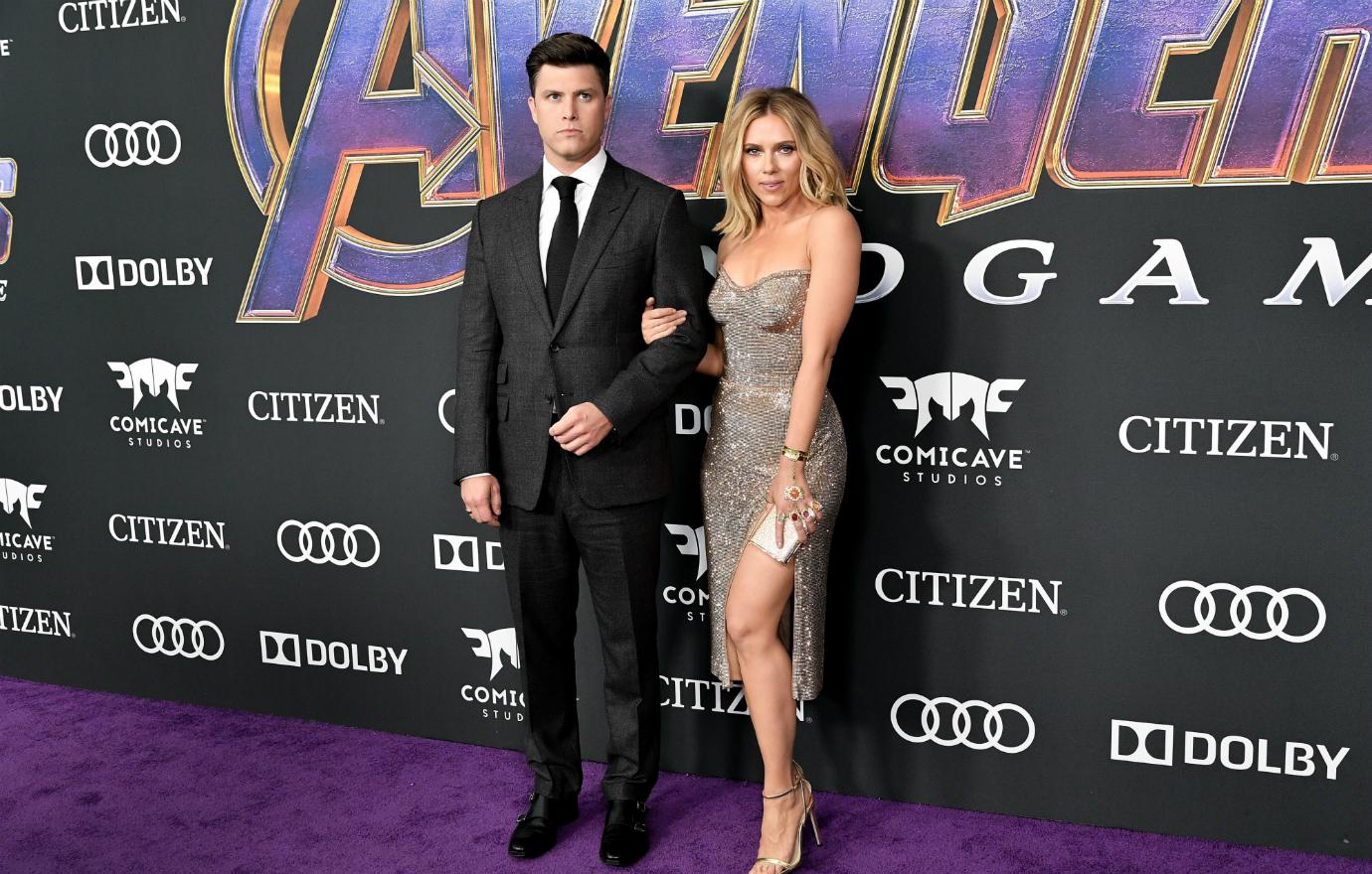 Scarlett Johansson, in a silver dress, stands with Colin Jost, in a black suit, on the red carpet.