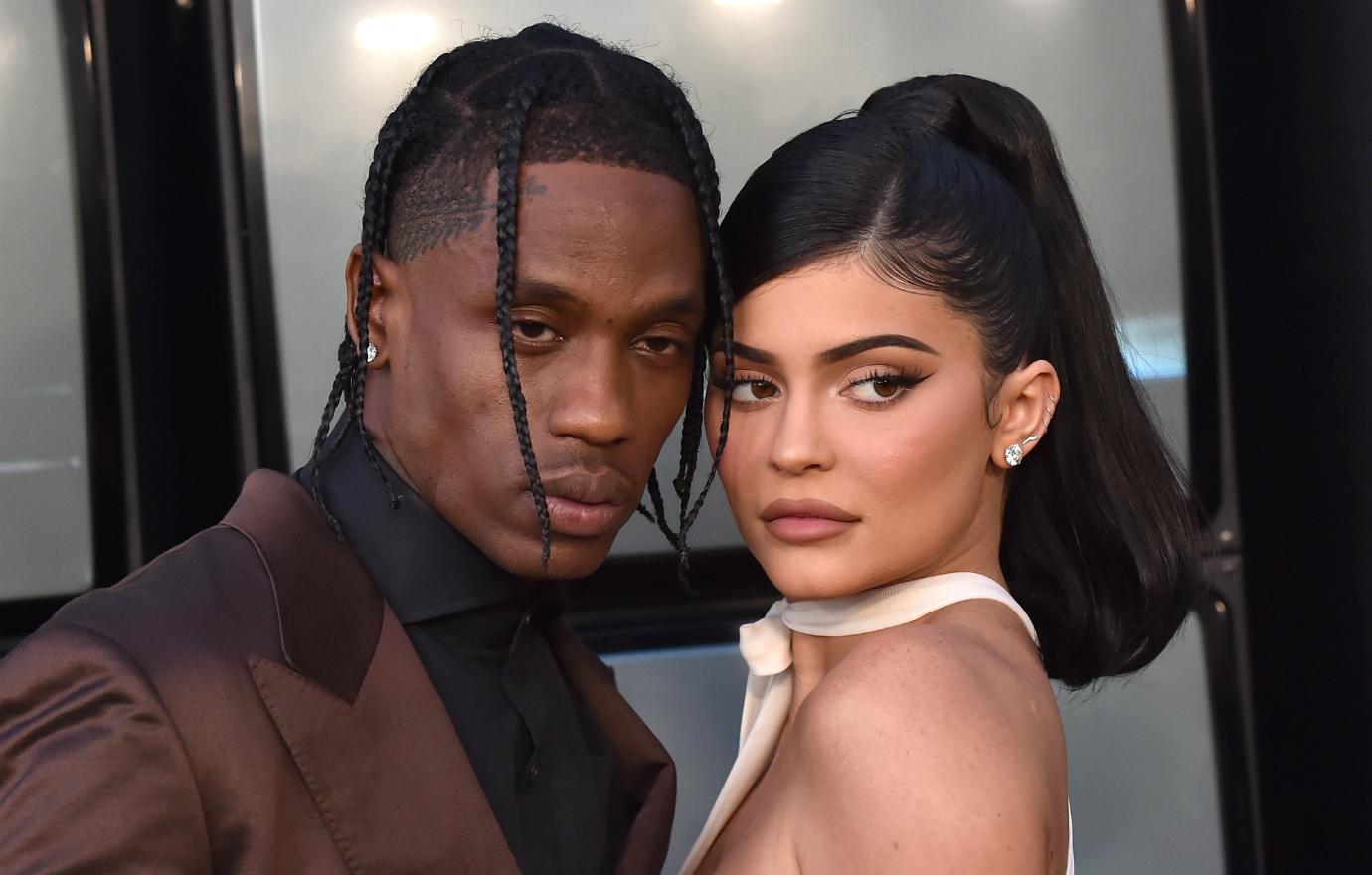 Travis Scott, in brown jacket and black shirt, rests his head on Kylie Jenner who wears a white dress.