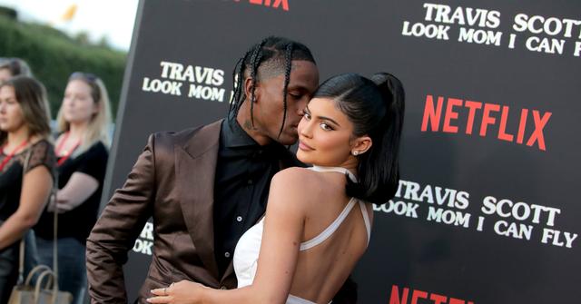 Travis Scott, in a brown jacket and black shirt, kisses Kylie Jenner, who wears a white dress, on the red carpet.