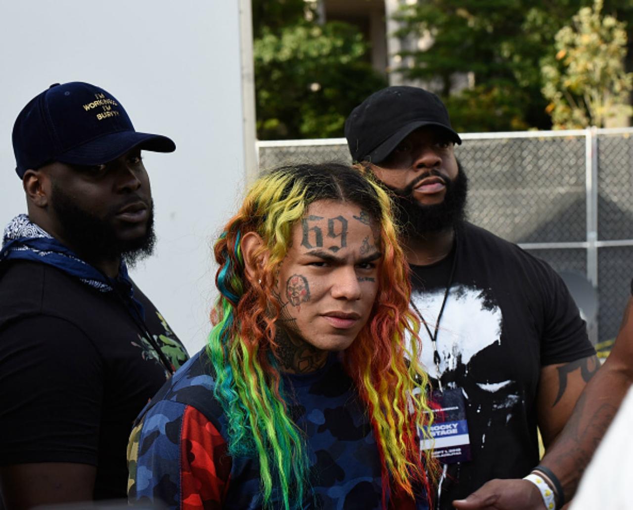 6ix9ine looks at the camera standing in front of two security guards.