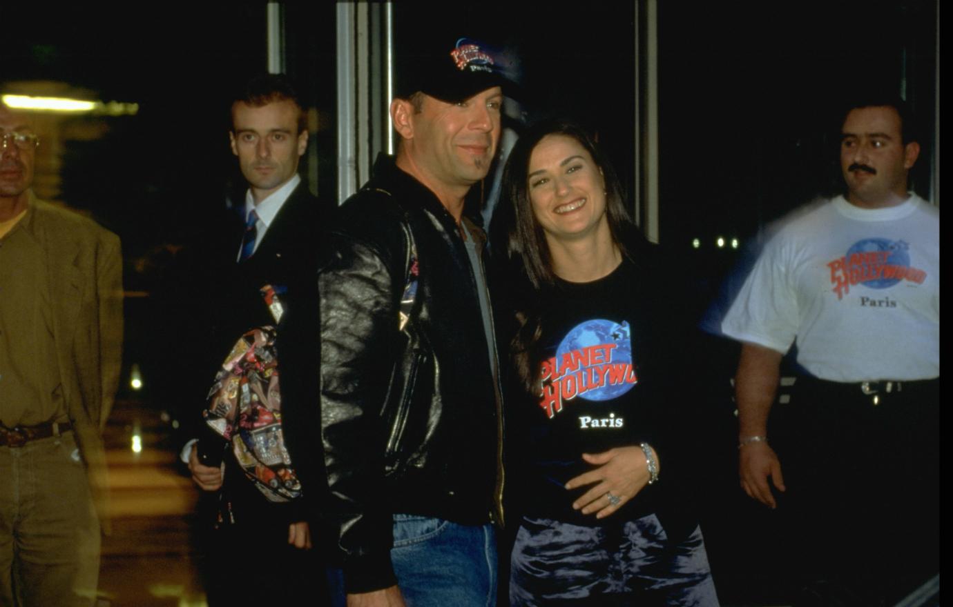 Bruce Willis in black jacket and black baseball cap stands next to Demi Moore who wears a Planet Hollywood Paris t-shirt.