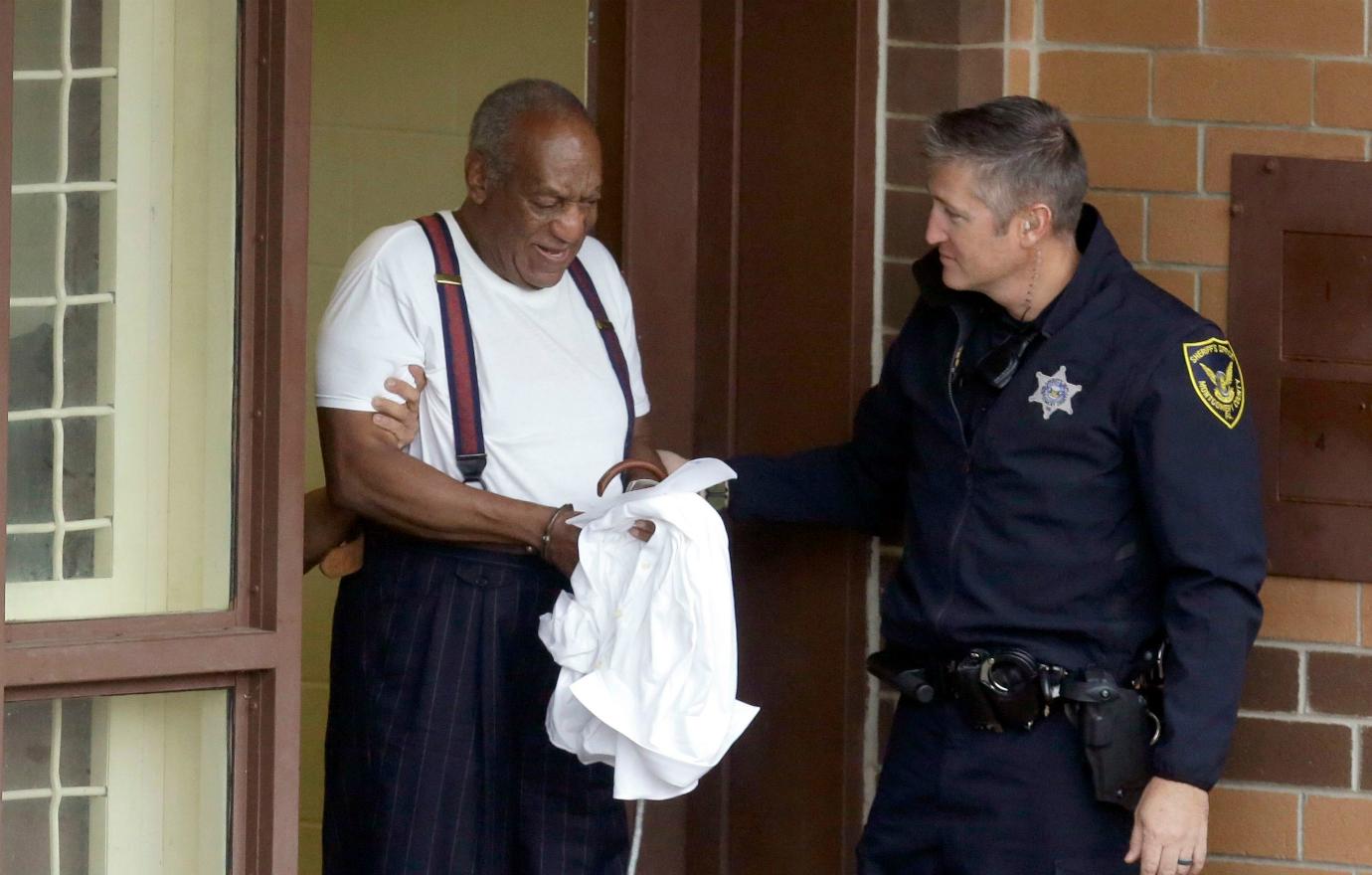 Bill Cosby, in a tshirt and suspenders, holds his shirt in his hands.