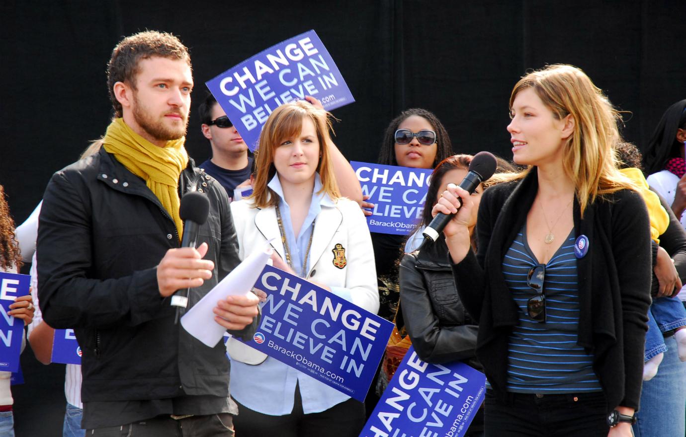 Justin Timberlake in black jacket and mustard-colored scarf holds a mic and a script while standing on stage alongside Jessica Biel who also holds a mic while wearing a blue striped top and a black jacket.