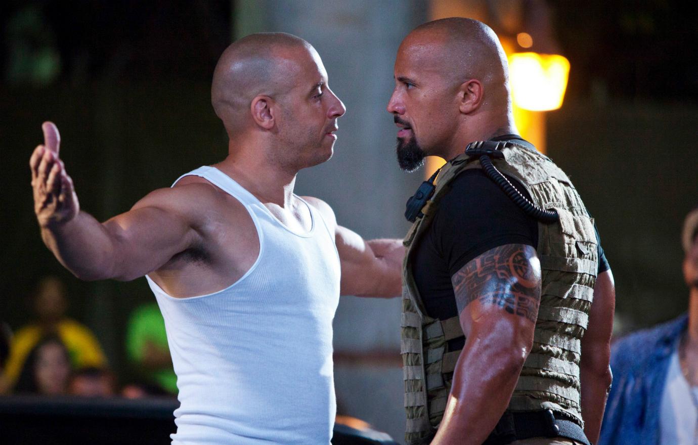 A scene from the Fast Five movie shows Vin Diesel, in a white tank top, stare off Dwayne Johnson who wears a flak jacket.