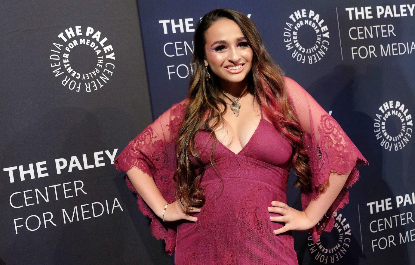 Jazz Jennings poses on the red carpet with her hands on her hips. She wears a maroon dress.