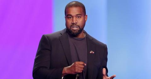 Kanye West, on stage and holding a mic, wears a dark jacket over a dark top.