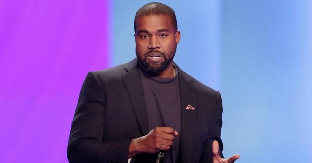 Kanye West, on stage and holding a mic, wears a dark jacket over a dark top.