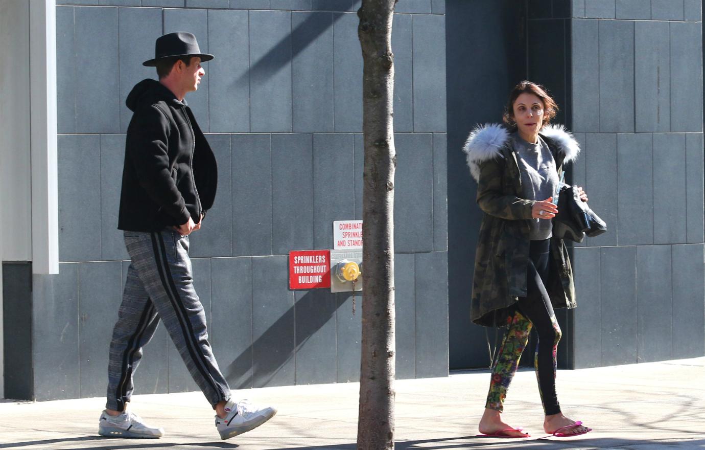 Paul Bernon, in dark hat, jacket, and grey pants walks up to Bethenny Frankel who wears a fur-lined jacket and leggings.