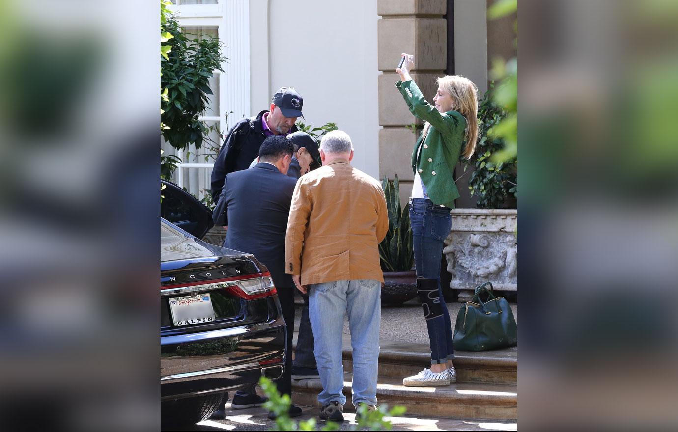 Larry King Wearing Baseball Cap Being Helped Out Of Car While Shawn Wearing a Green Jacket And Jeans Takes Photos of Trees With Phone