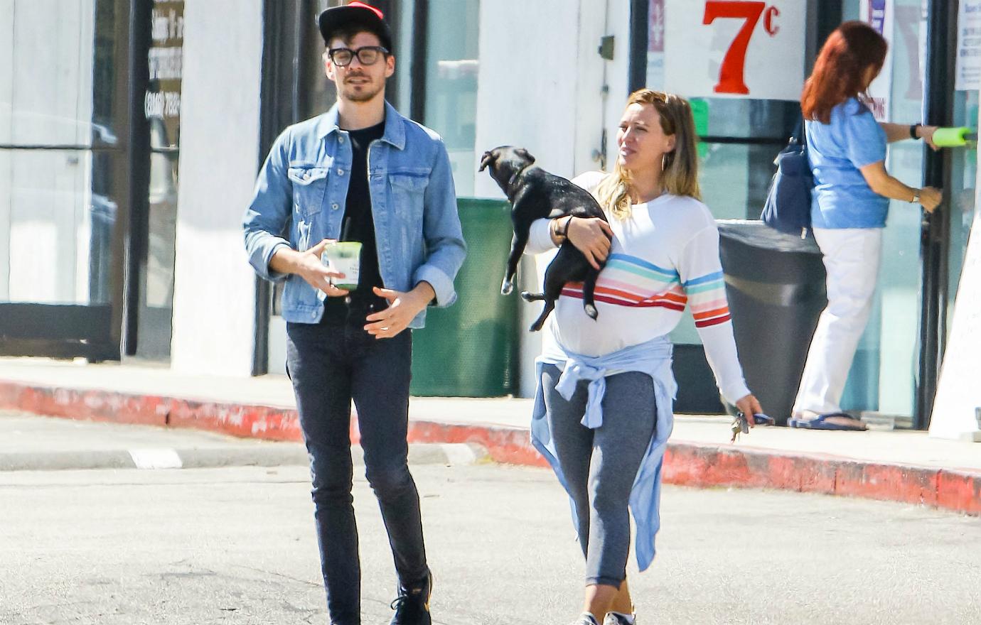 Hilary Duff held Lucy the dog with a white rainbow-adorned sweater and grey yoga pants while Matt Koma wore a jean jacket, black T-shirt and dark pants.