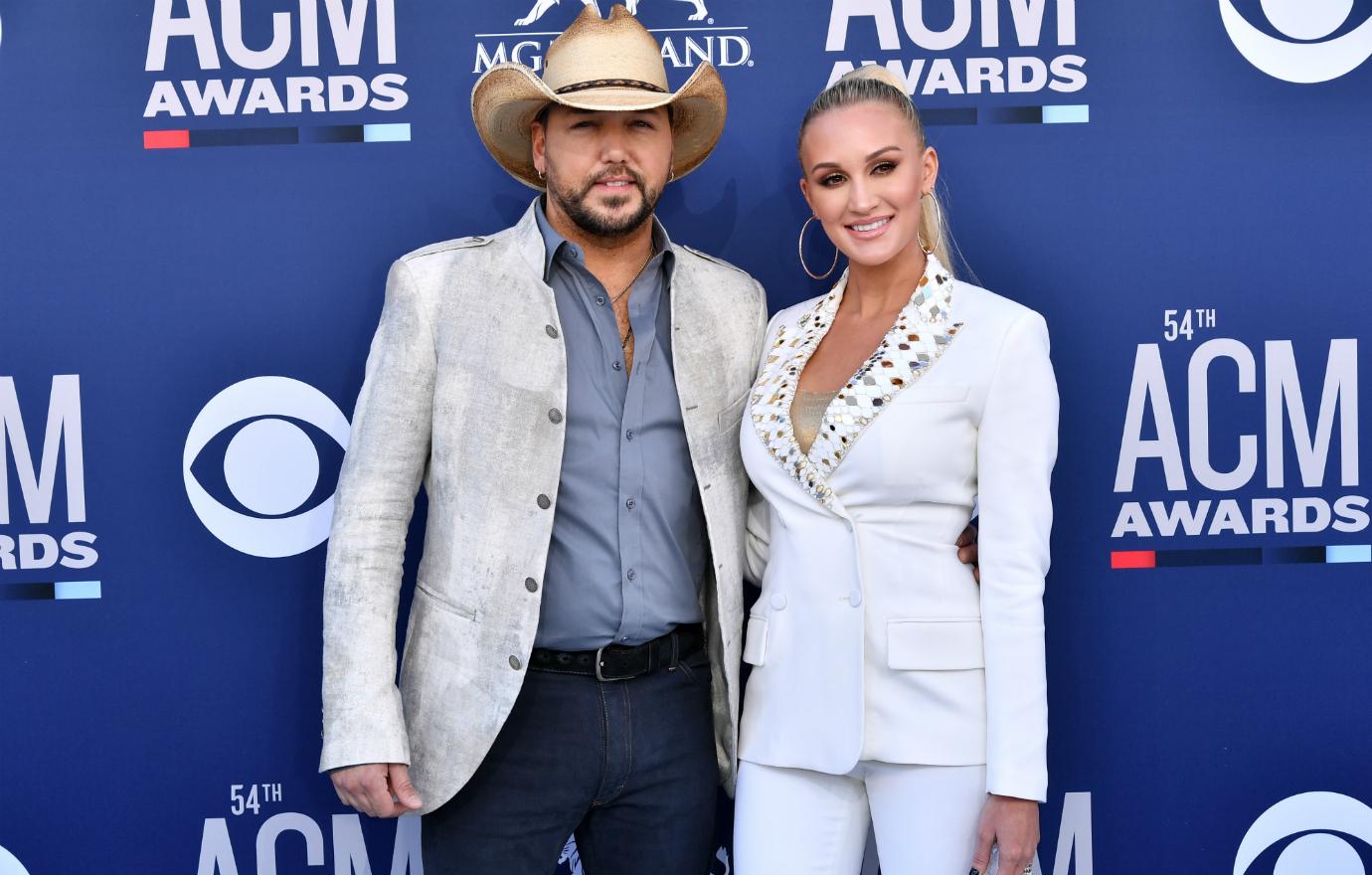 Jason Aldean, in cowboy hat, jacket and jeans, poses alongside Brittany Kerr who wears a white pant suit with golden embellishments.