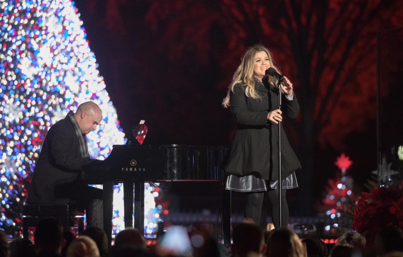 Kelly Clarkson wearing a black dress, singing in front of a Christmas Tree