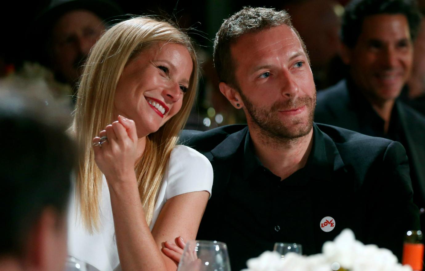 Gwyneth Paltrow, in a white top, sits at a table, alongside Chris Martin who wears a black suit and black shirt.