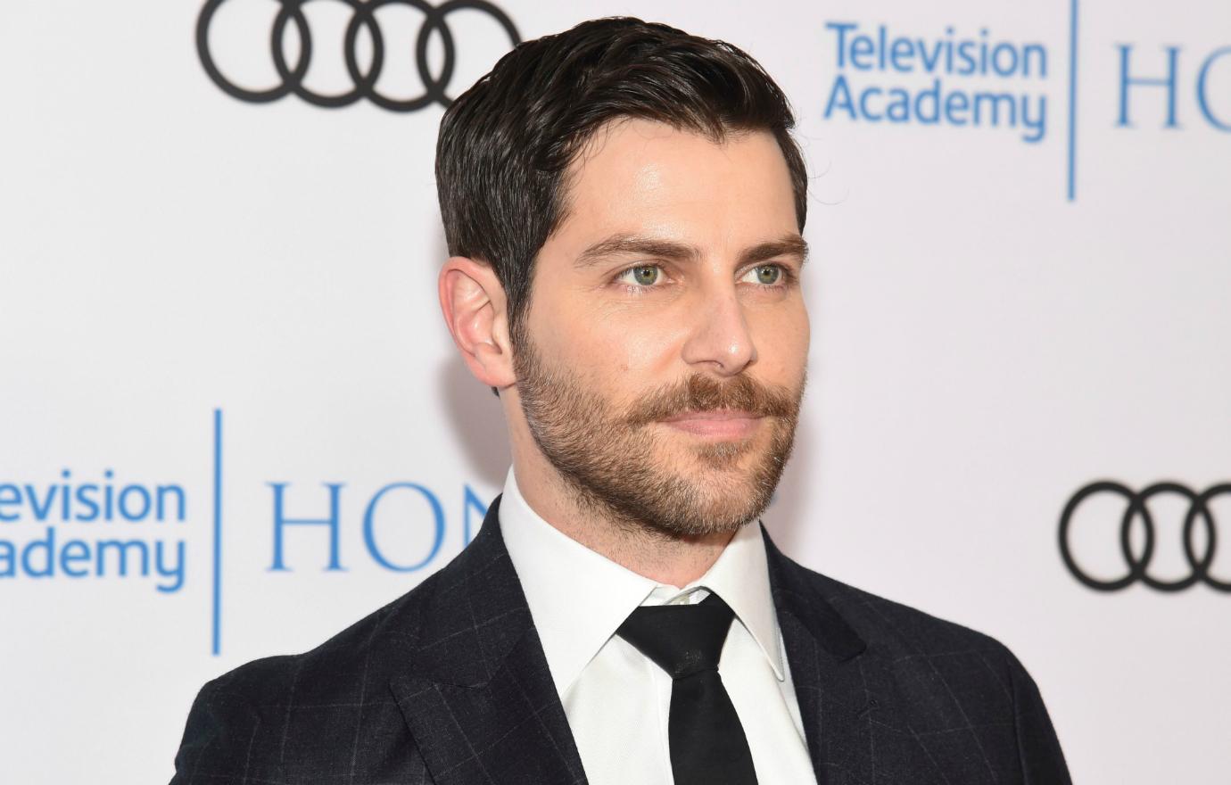 David Giuntoli in a black suit and tie and white shirt stands on the red carpet.