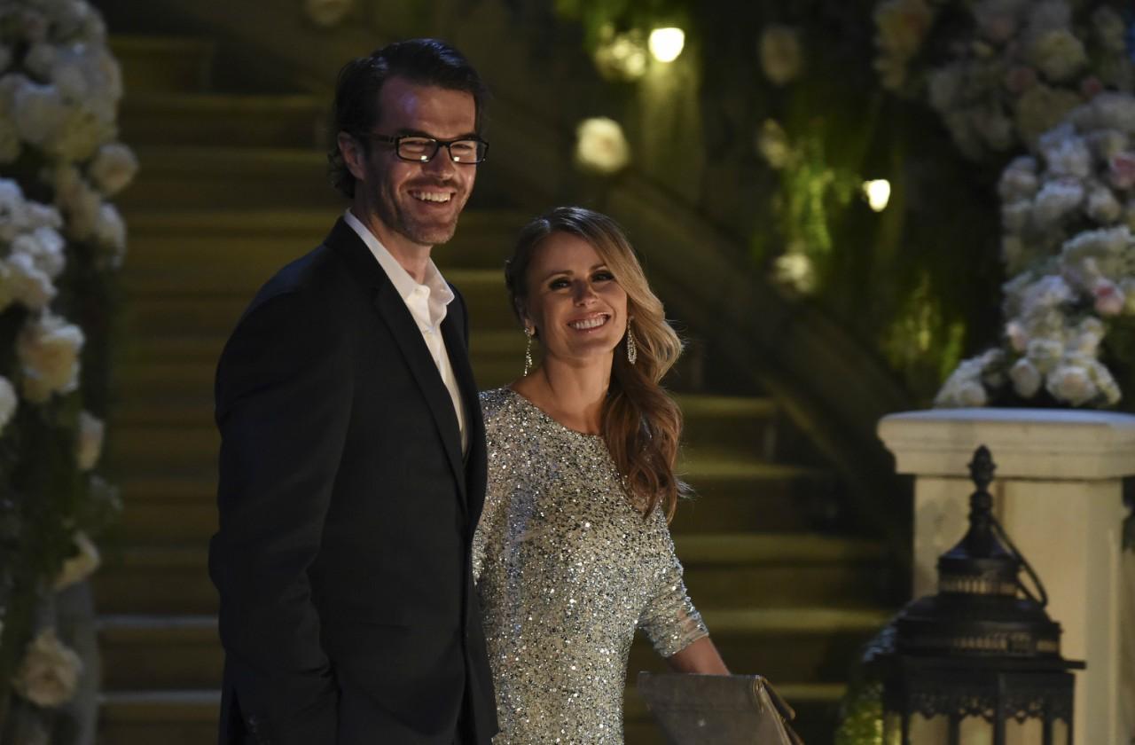 Ryan Sutter in a black suit stands in front of bachelor mansion with wife, Trista, a white gown