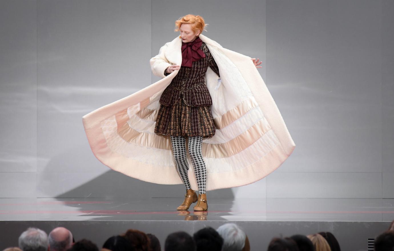 Tilda Swinton stands on stage in a big, twirling white coat.